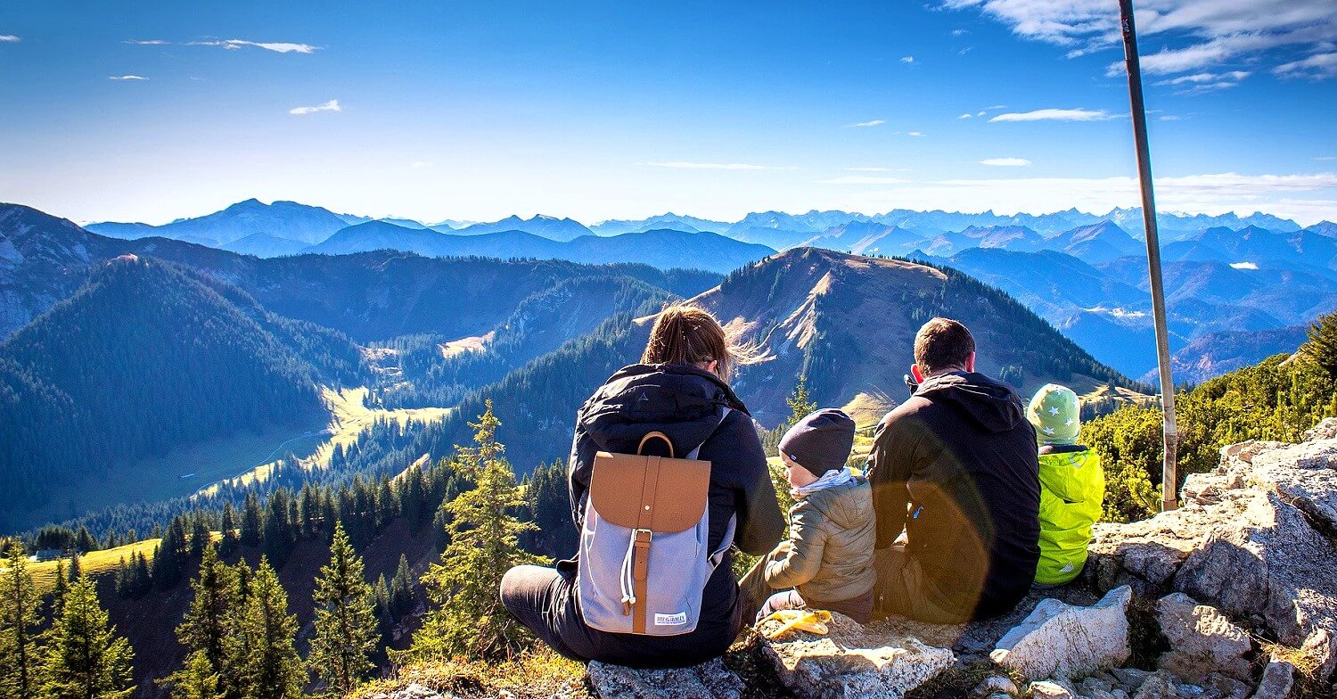 Eine Familie mit Rucksäcken rastet auf einem Berggipfel und genießt die Aussicht über die Landschaft aus Bergen und Wäldern bei strahlend blauem Himmel