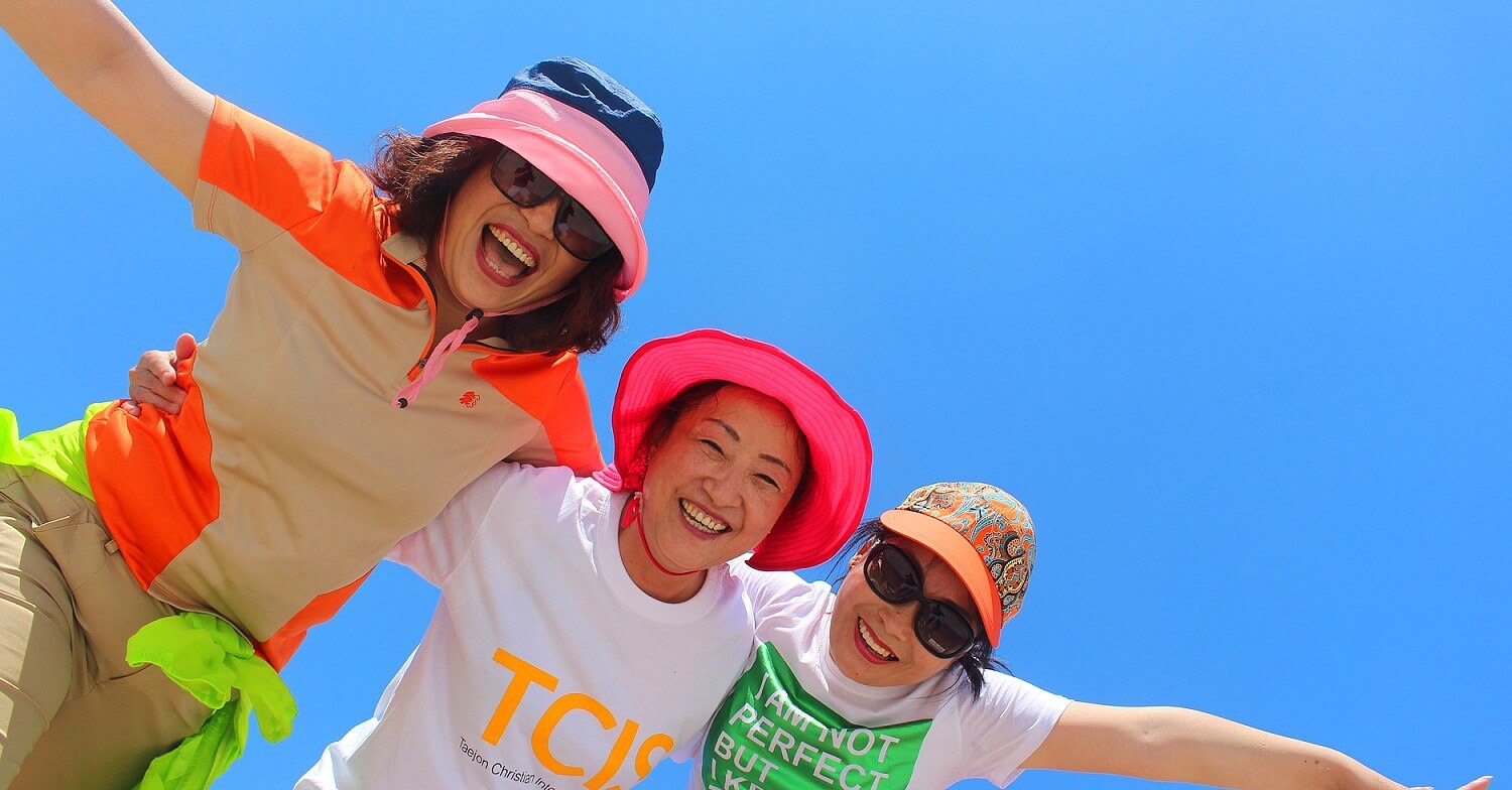 Drei lachende Frauen, die sich umarmen. Alle Frauen tragen T-shirts und einen Sonnenhut