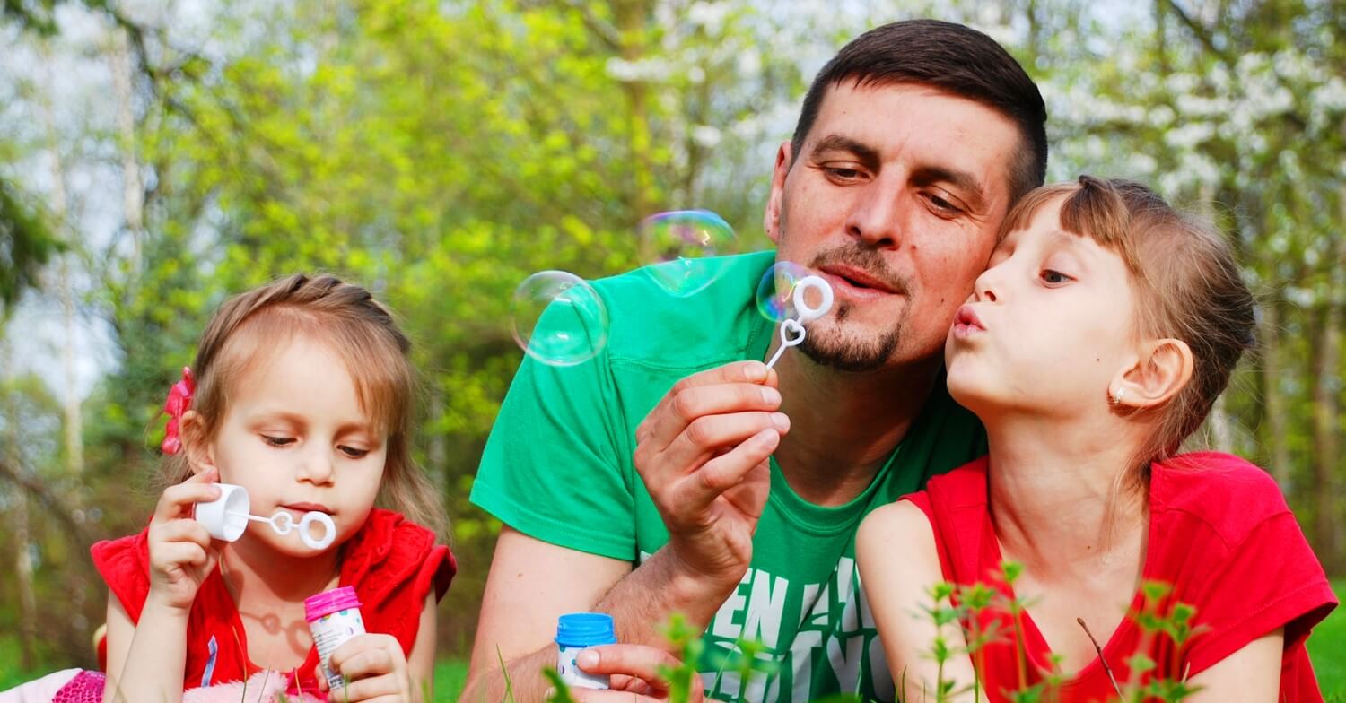Vater mit grünem T-shirt und seine beiden Töchter mit roten Shirts machen Seifenblasen