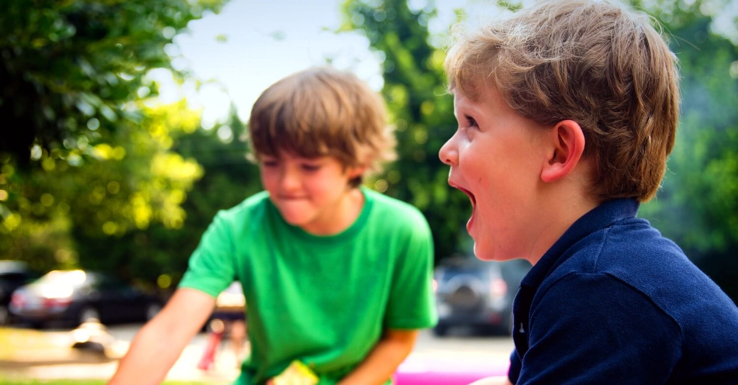 Zwei Jungen sitzen im Sandkasten. Ein Junge trägt ein grünes T-Shirt und spielt im Sand, der andere Junge trägt ein blaues Oberteil und lacht