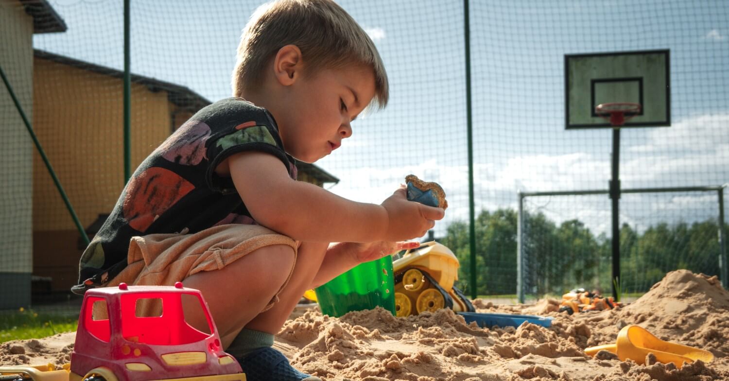 Zwei Jungen sitzen im Sandkasten. Ein Junge trägt ein grünes T-Shirt und spielt im Sand, der andere Junge trägt ein blaues Oberteil und lacht