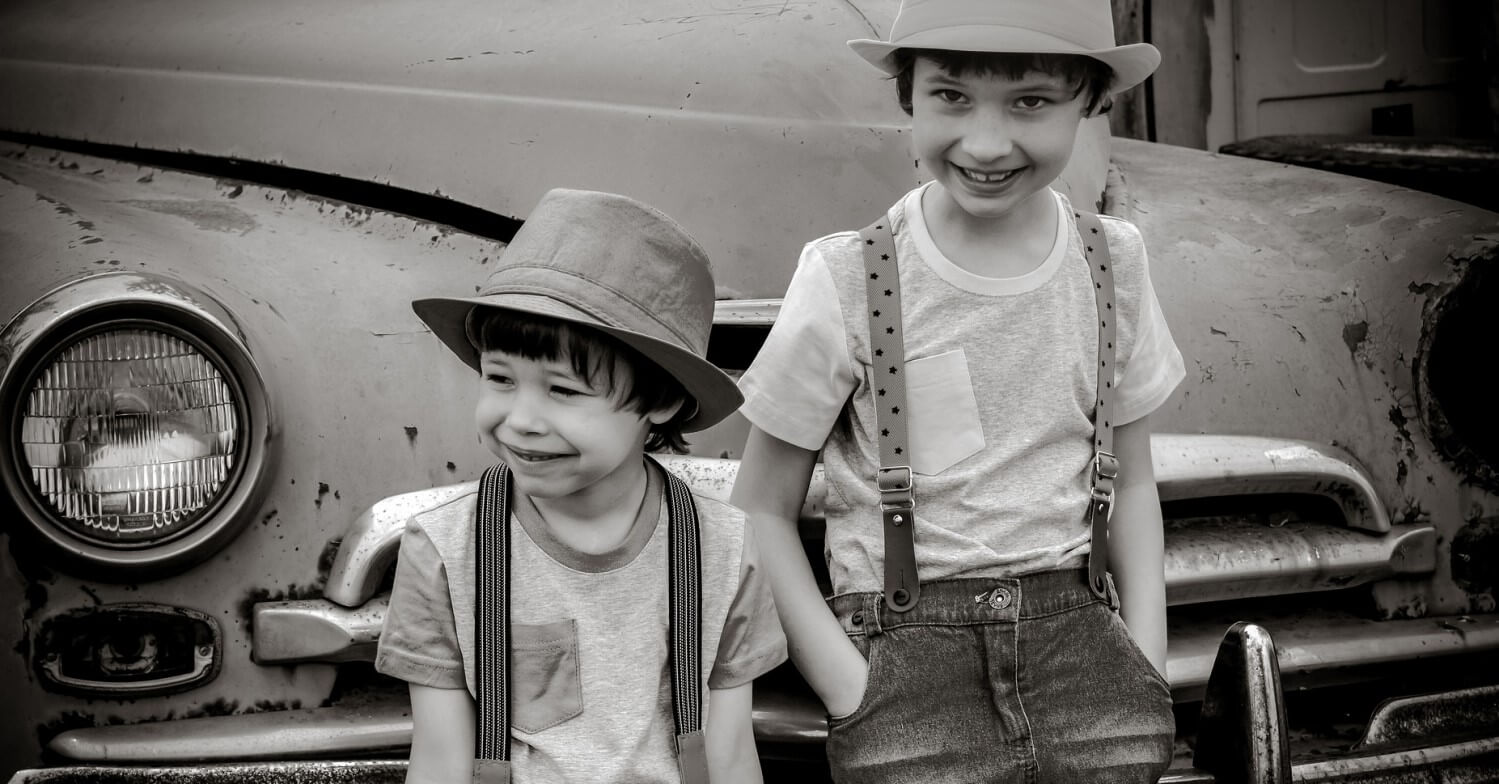 Schwarzweißfoto von zwei kleinen Jungen mit Hosenträgern und Hut vor einem Auto