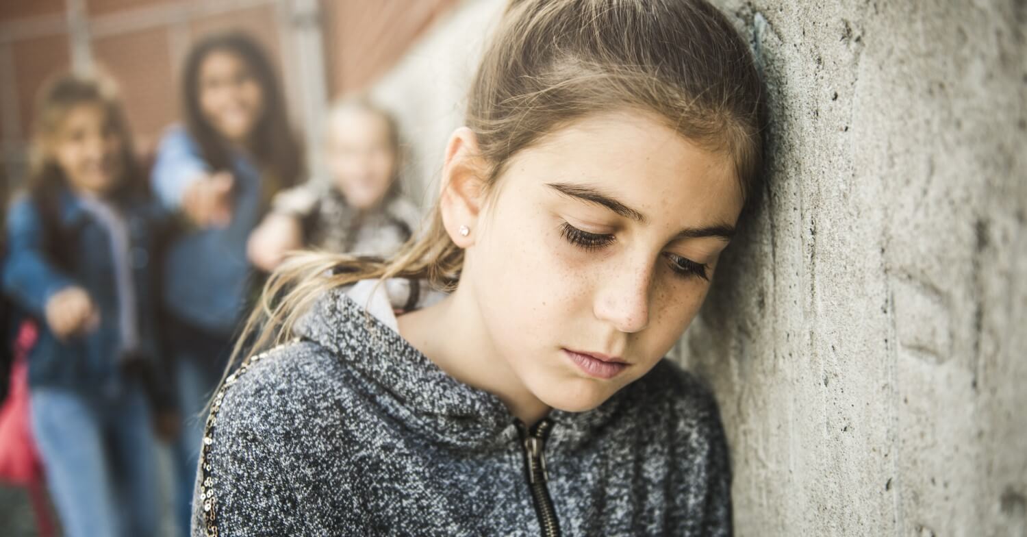 Ein Mädchen lehnt gegen eine Wand und schaut traurig. Im Hintergrund stehen mehrere Schülerinnen, die mit dem Finger auf die zeigen
