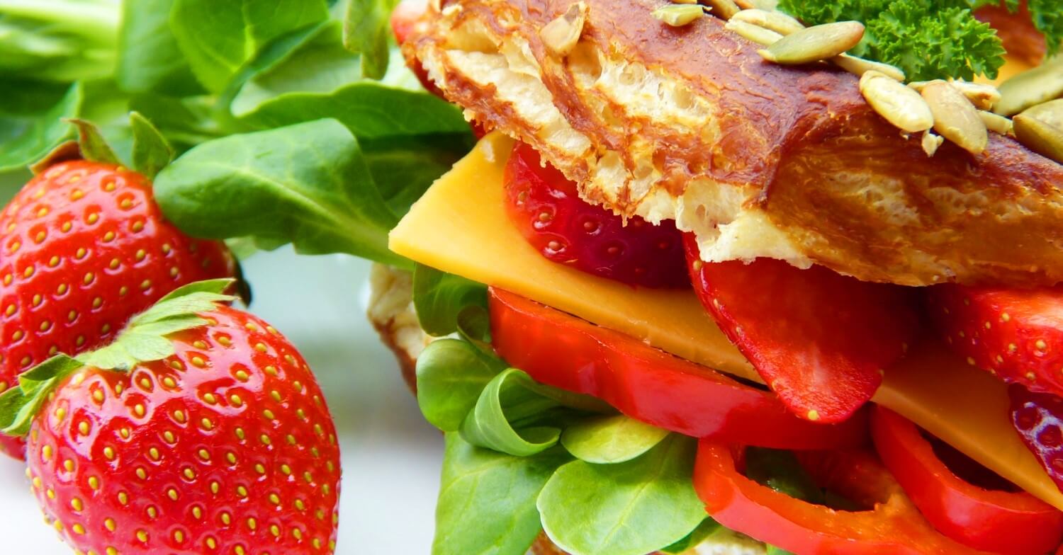 Ein gesundes Schulbrot: Sandwich mit Gemüse, Salat und Käse belegt, dazu ein paar Erdbeeren