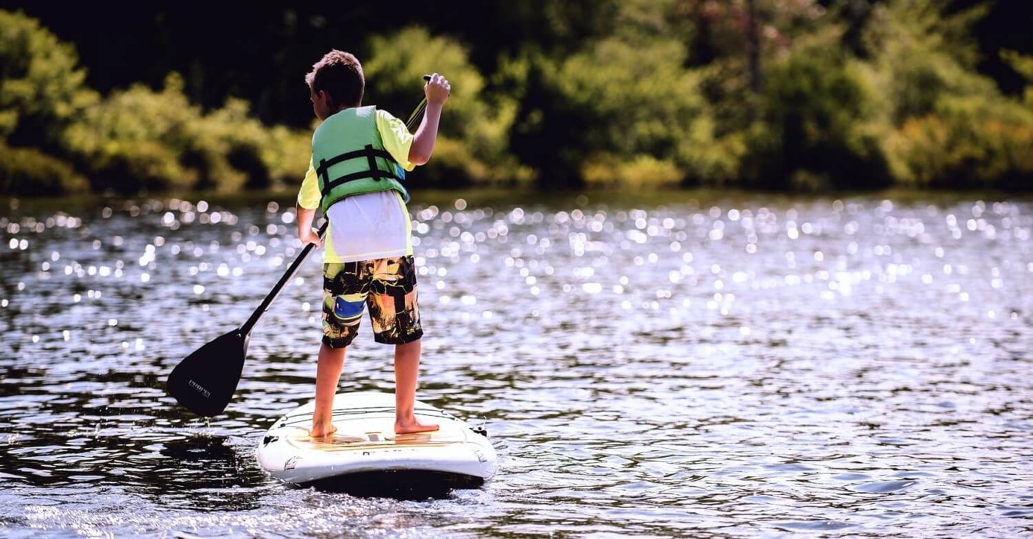 Ein Junge steht mit einem Paddel auf einem Surfbrett und paddelt über einen See